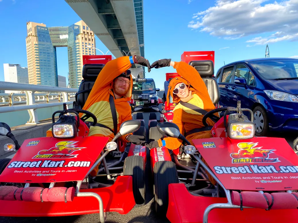 Two people in orange mascot outfits sit in red go‑karts labeled Street Kart, forming a heart with their arms in front of a city bridge.