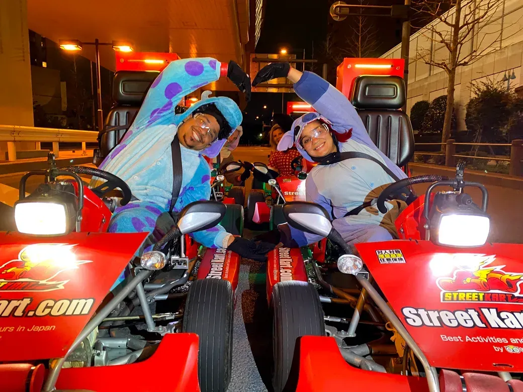 Two people in colorful onesies pose in red Street Kart go-karts at night, forming a heart shape with their arms.