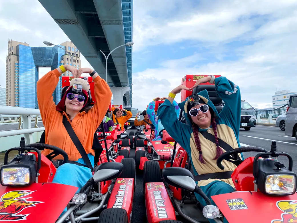 Group of people in red go-karts under a bridge, making heart shapes with their arms and wearing colorful outfits and sunglasses. A fun, communal activity on a city street race track.